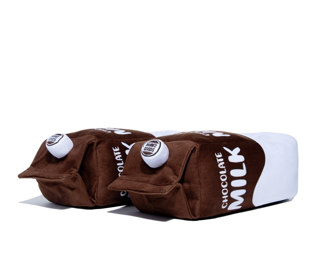 A novelty plush slippers shaped like two chocolate milk cartons side by side, featuring brown and white colors with 'CHOCOLATE MILK' printed in white text, set against a plain white background.