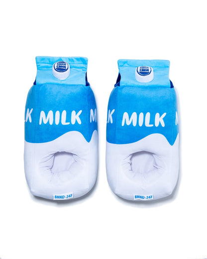 Two plush milk carton-shaped slippers displayed side-by-side on a clean white background. The slippers are designed to look like milk cartons, featuring a bright blue top half with white 'MILK' lettering and a white bottom half.