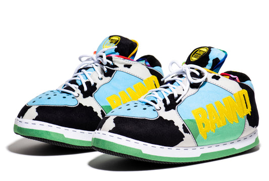 A pair of low-top sneakers with a cow-print pattern in black and white, light blue suede overlays, a mint green heel section, and bold yellow 'BANND' lettering on the side. The shoes are photographed on a clean white background with a soft shadow.